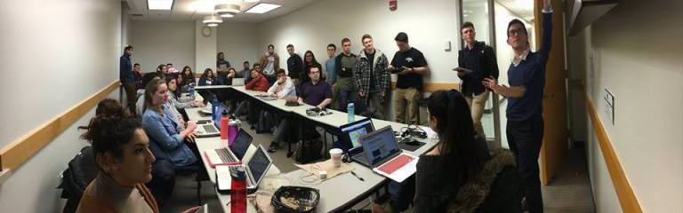 Students crowded in a small room with tables and laptops in the center